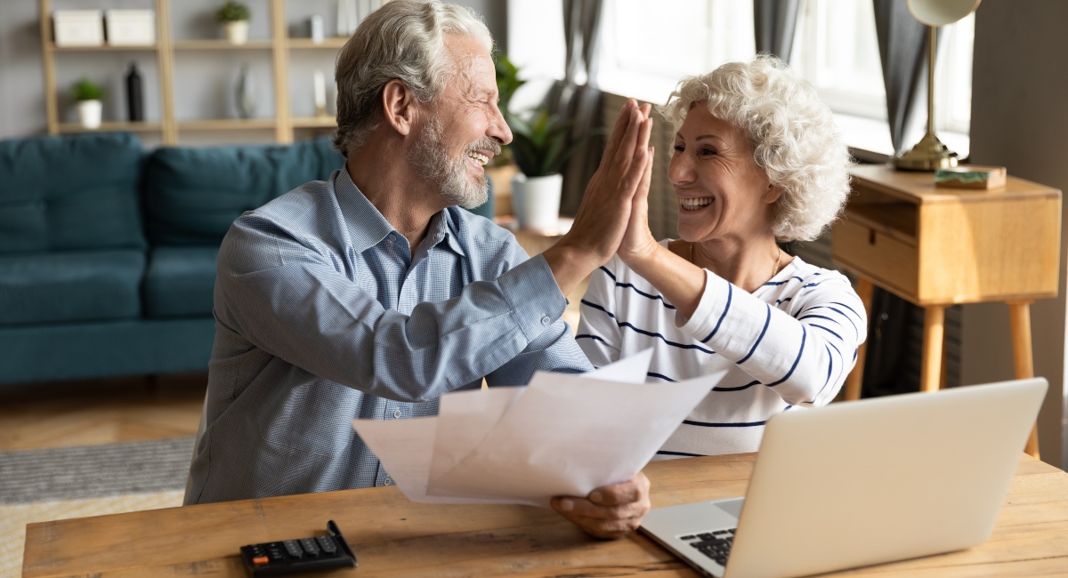 Older couple giving each other a "high-five" sitting at a desk with a laptop on it