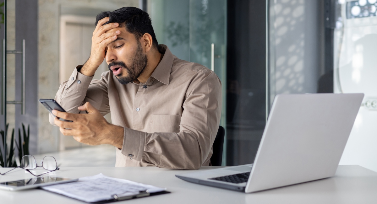 man sitting at desk looking at phone with worried expression