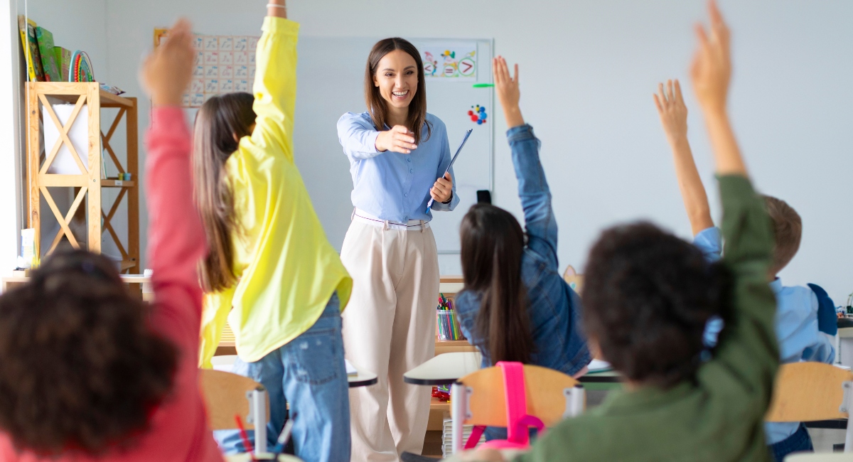teacher wearing blue shirt standing in front of classroom with kids who have their hands raised