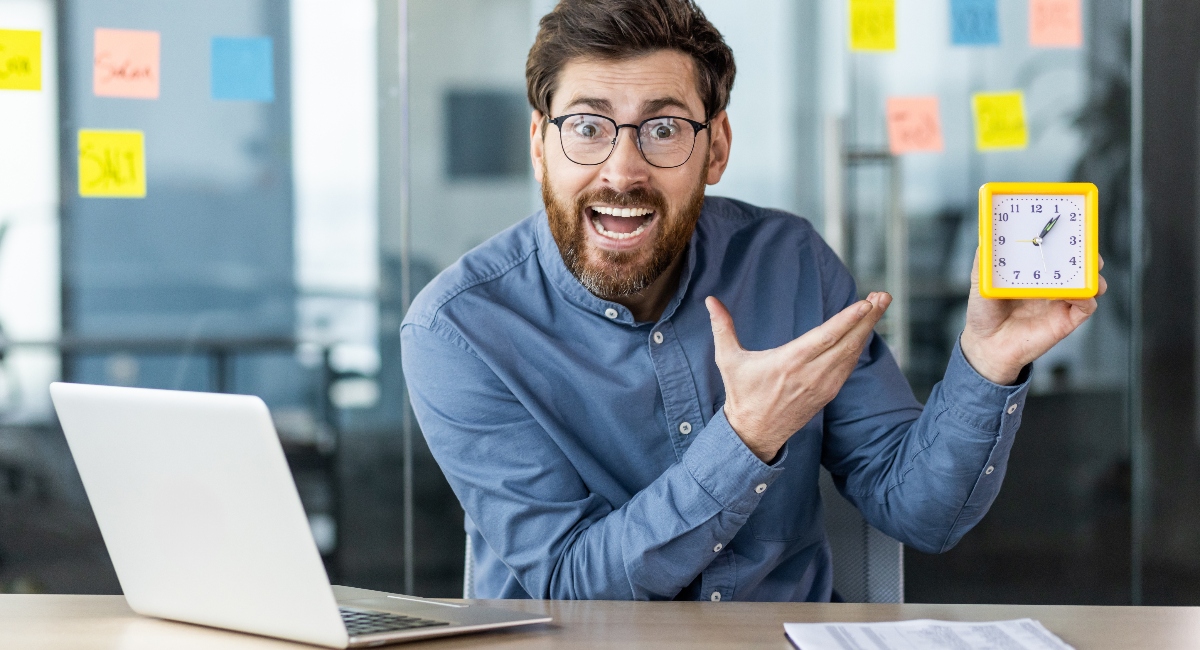 Worried businessman sitting in the office at the desk, pointing at the alarm clock in his hand.