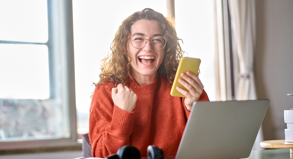 Young professional sitting at computer holding a yellow phone in her hand, smiling and excited