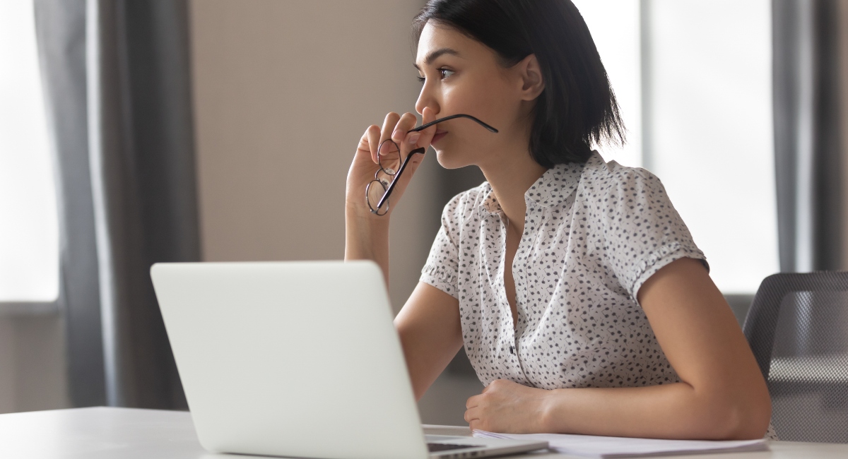 worried woman sitting at a desk holding eyeglasses in her hand, gazing into the distance