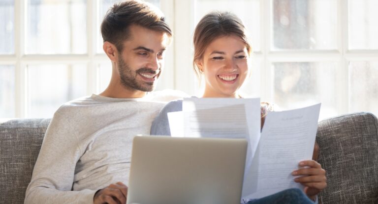 Smiling husband and wife sit on couch using laptop