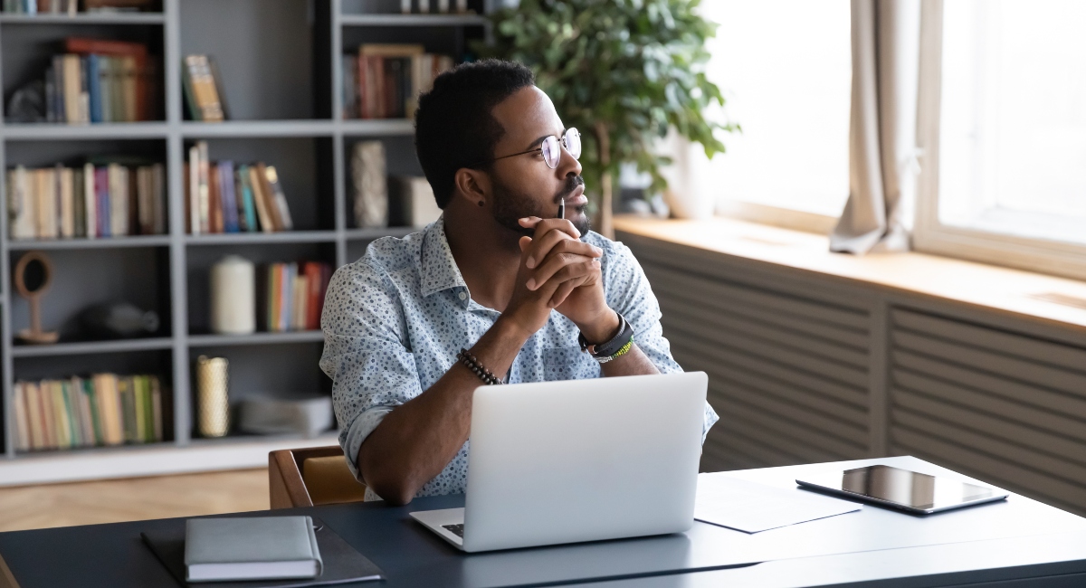 Thoughtful businessman sitting at table with computer, looking away and out the window