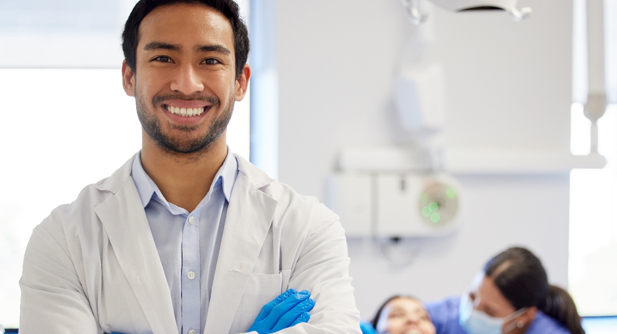 Male dentist in white coat standing in a dental exam room with a patient in the chair in the background